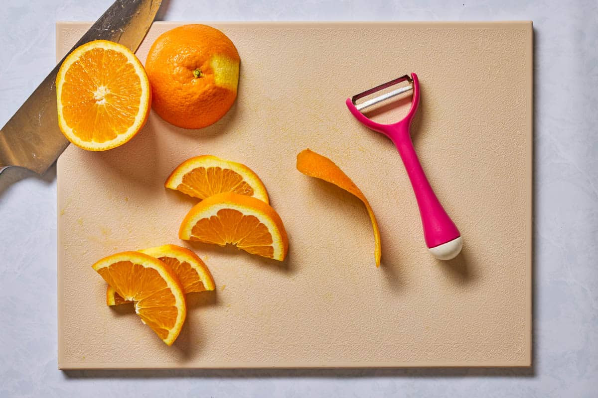 Orange halves, slices, and a peel on a cutting board with a knife and a peeler.