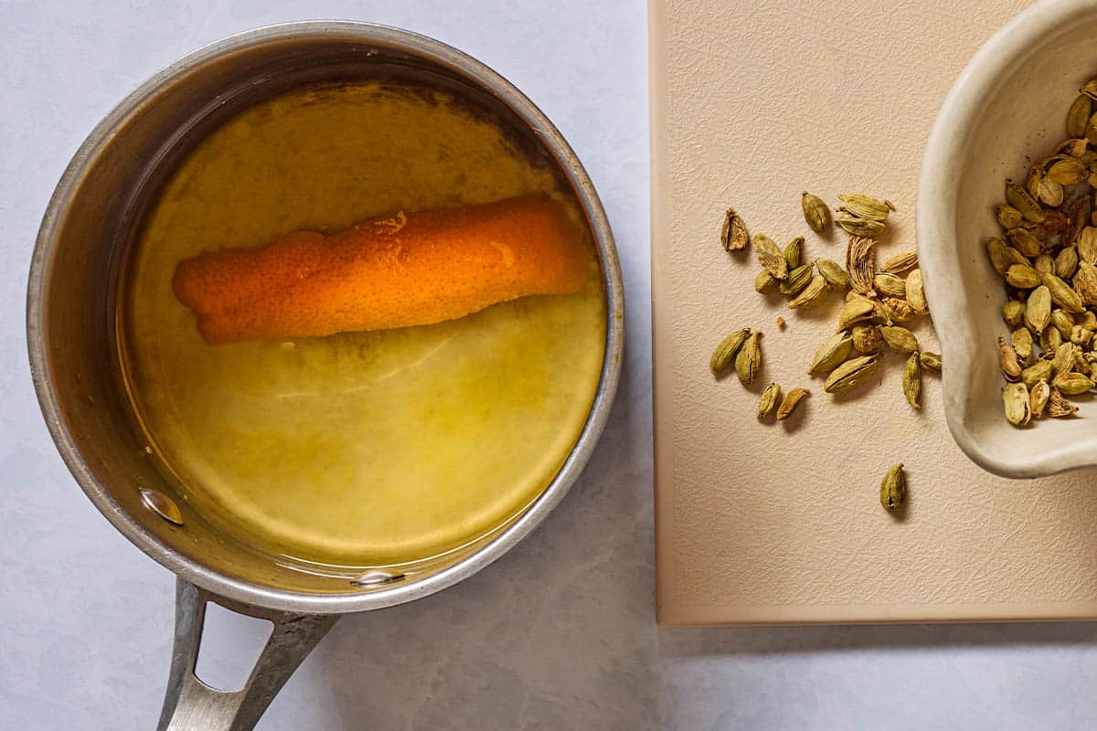 The syrup for the mocktail in a saucepan with an orange peel. Next to this are some cardamom seeds on a cutting board and in a bowl.