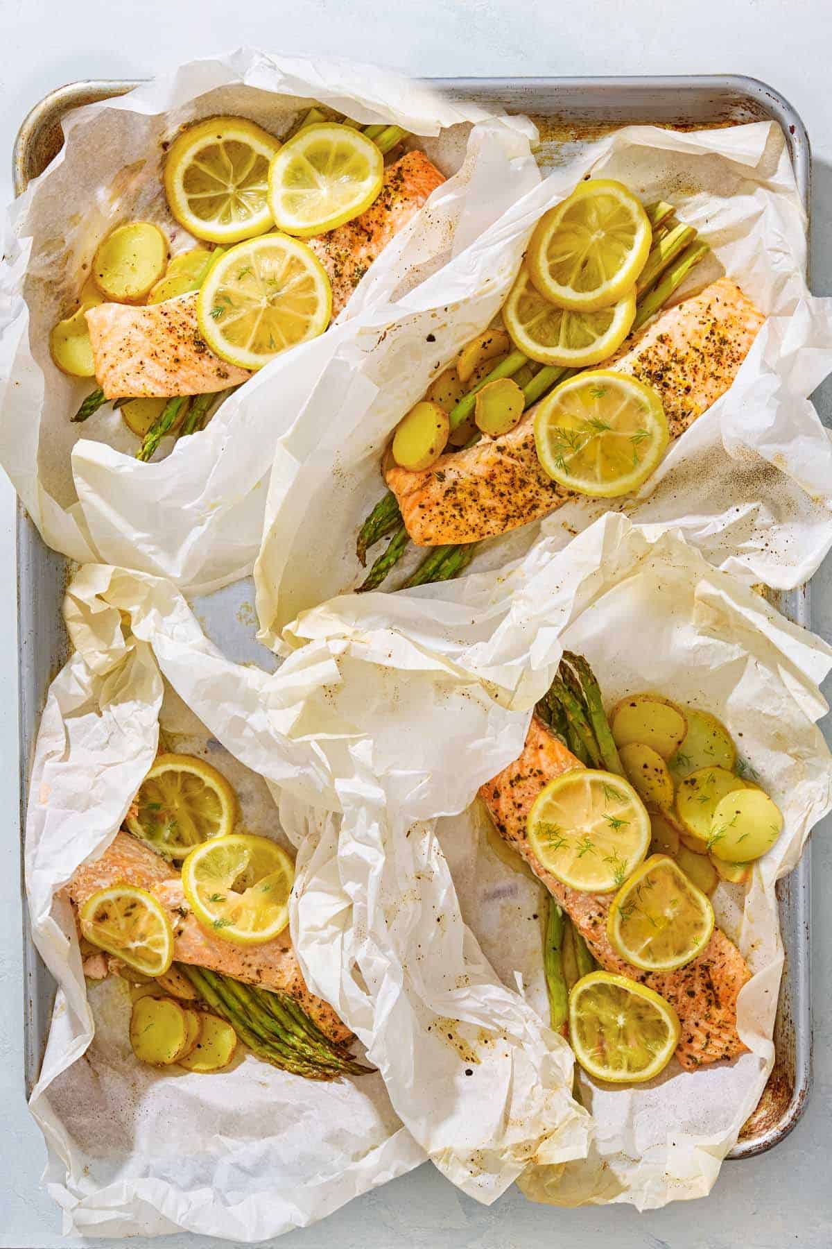 4 open salmon en papillote topped with lemon wheels on beds of asparagus and potatoes on a baking sheet.