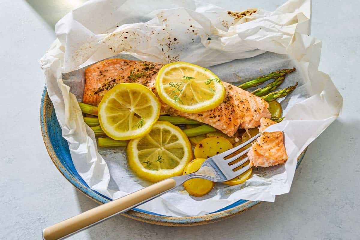 Salmon en papillote topped with lemon wheels on a bed of asparagus and potatoes on a plate with a fork.