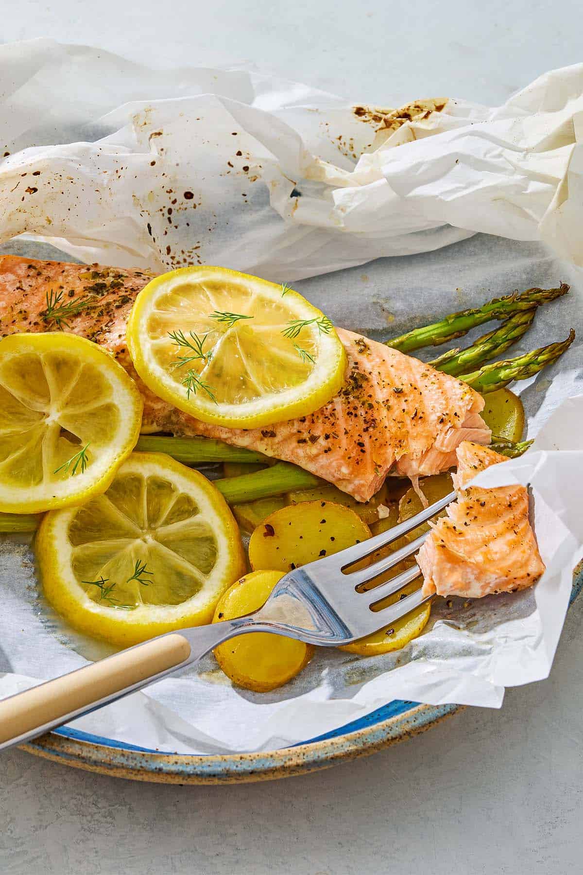 Salmon en papillote topped with lemon wheels on a bed of asparagus and potatoes on a plate with a fork.