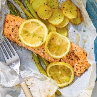Salmon en papillote topped with lemon wheels on a bed of asparagus and potatoes on a plate with a fork.
