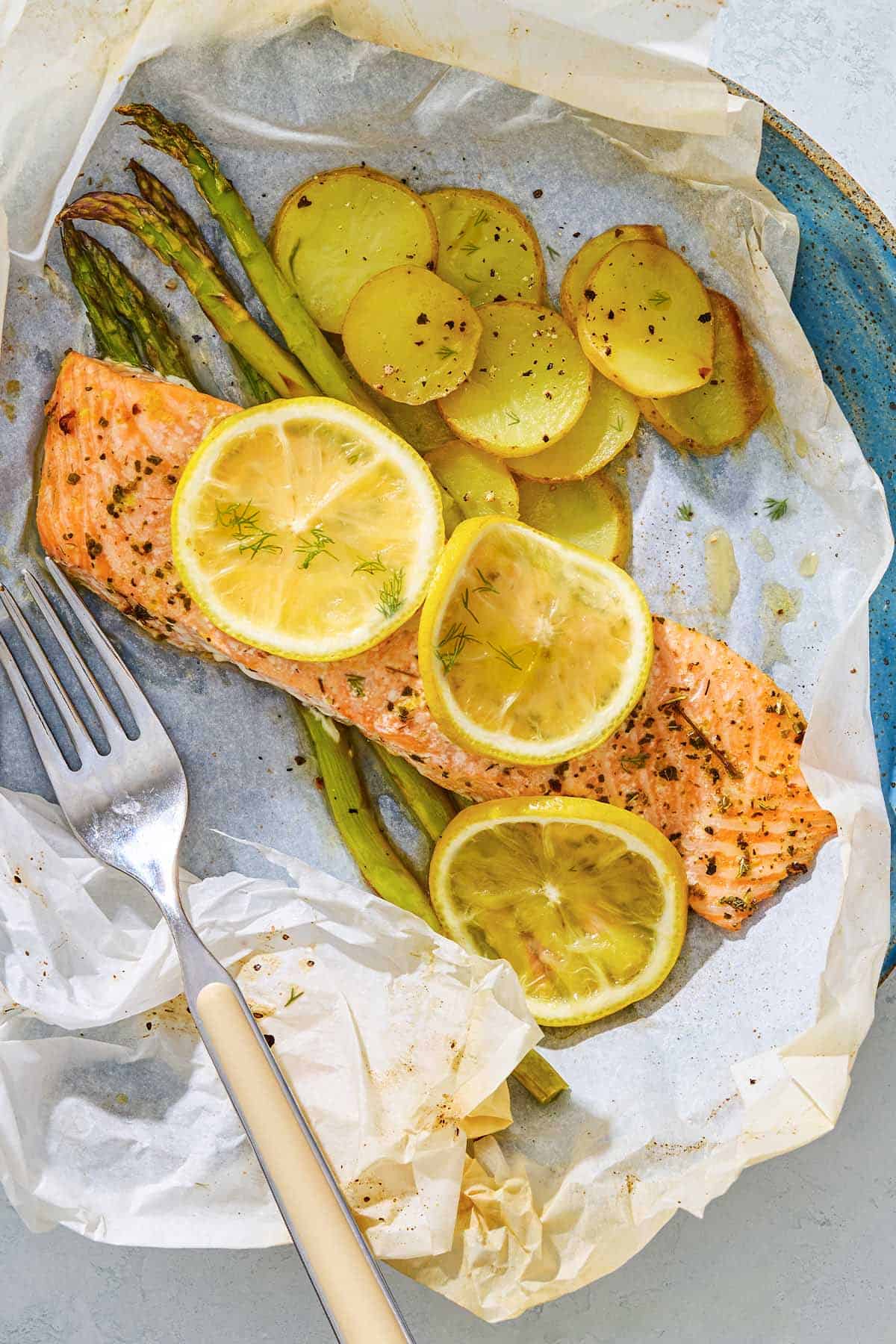 Salmon en papillote topped with lemon wheels on a bed of asparagus and potatoes on a plate with a fork.