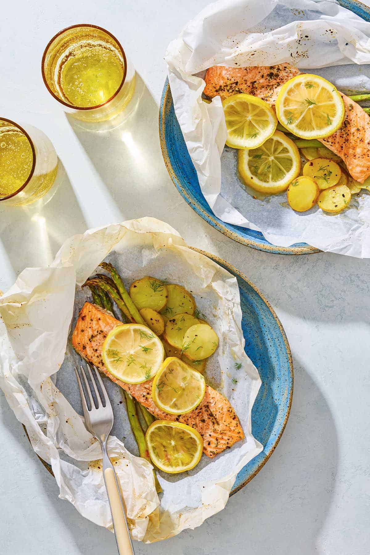 Two plates of salmon en papillote topped with lemon wheels on beds of asparagus and potatoes, one with a fork. Next to this are two cups of water.