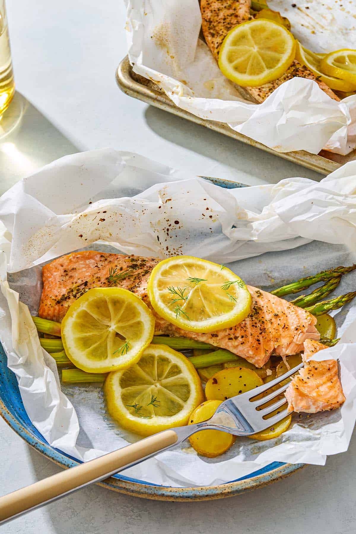 Salmon en papillote topped with lemon wheels on a bed of asparagus and potatoes on a plate with a fork next to the baking sheet with the rest of the salmon.