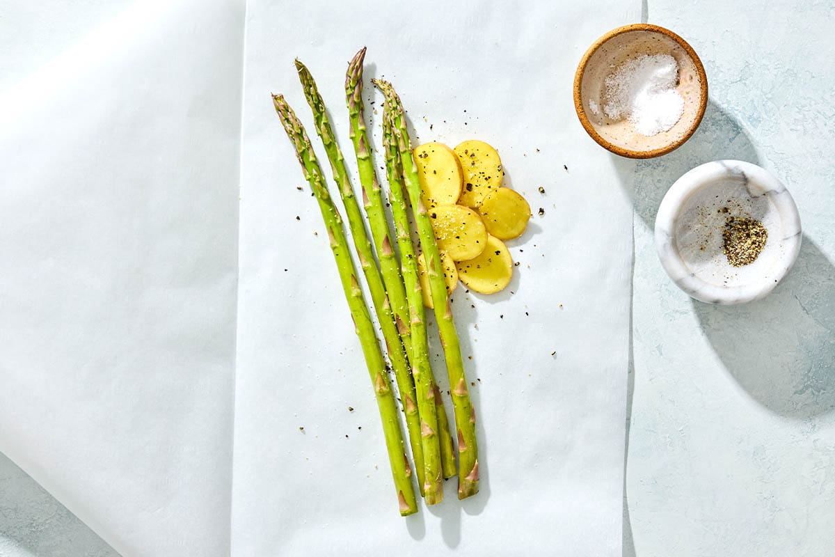 Unbaked asparagus and potatoes sitting on a sheet of parchment paper next to small bowls of salt and pepper.