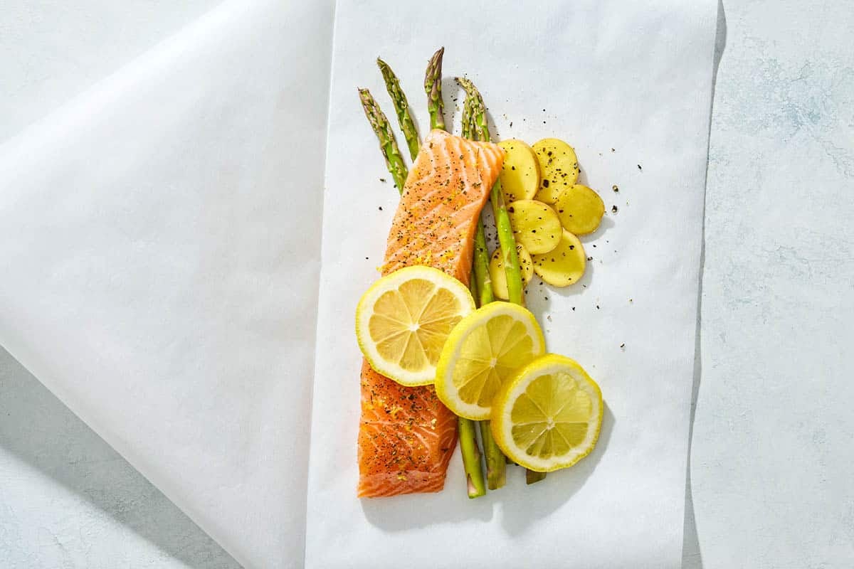 Unbaked salmon topped with lemon wheels on a bed of asparagus and potatoes sitting on a sheet of parchment paper.