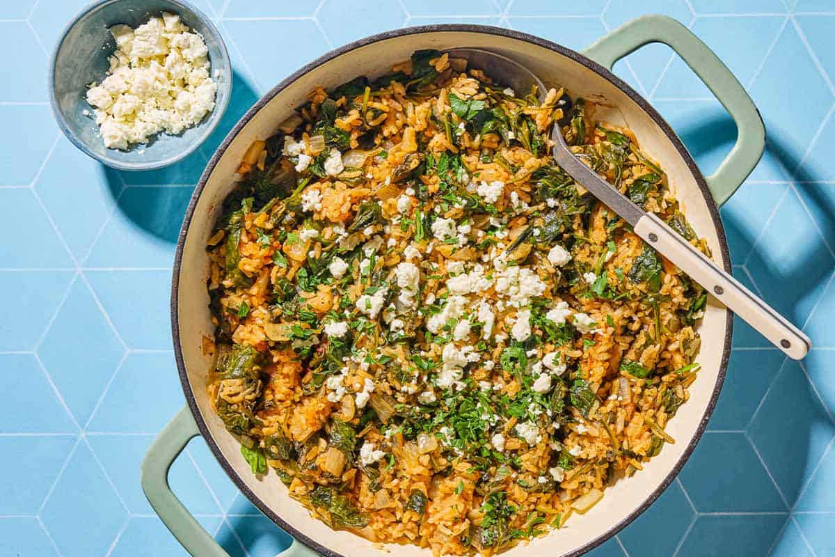 An overhead photo of spanakorizio (Greek spinach rice recipe) in a skillet with a serving spoon next to a bowl of feta.