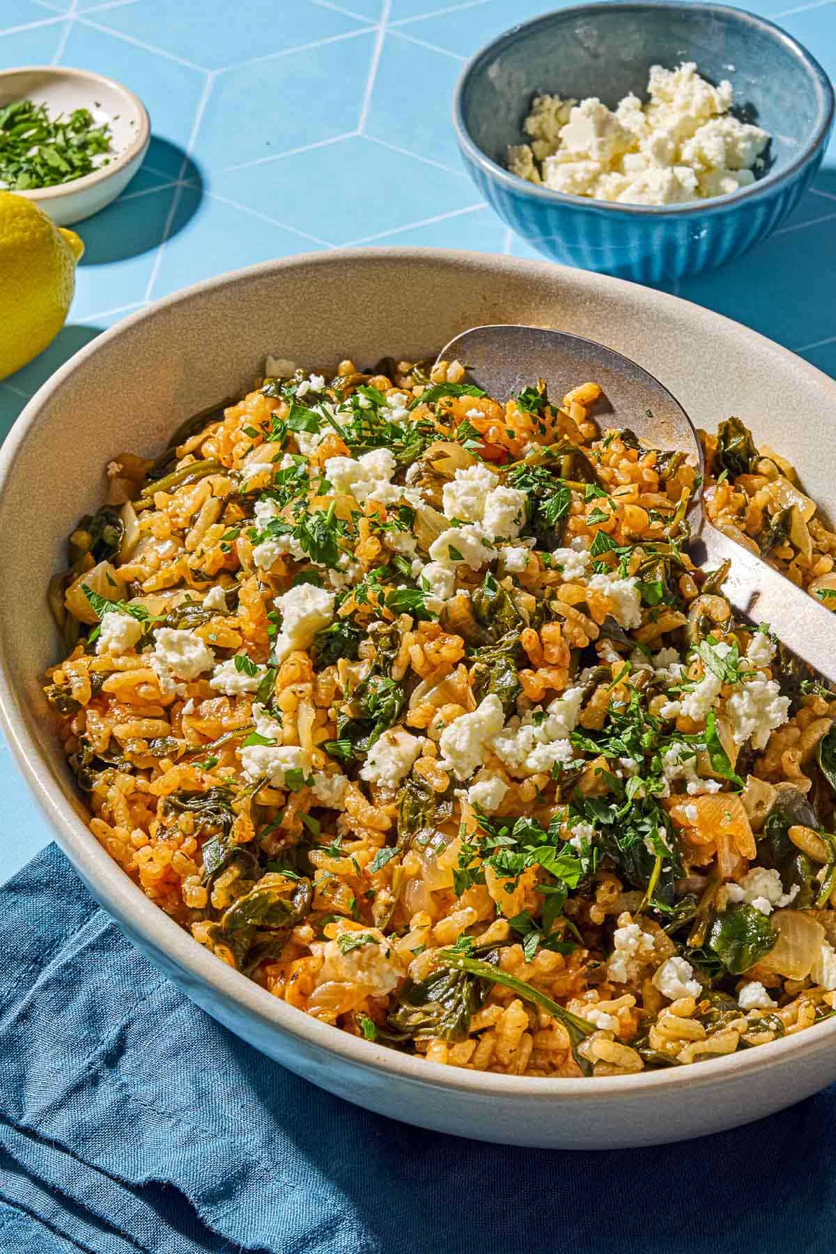 Spanakorizo in a serving bowl with a serving spoon surrounded by a lemon and bowls of feta and chopped parsley.
