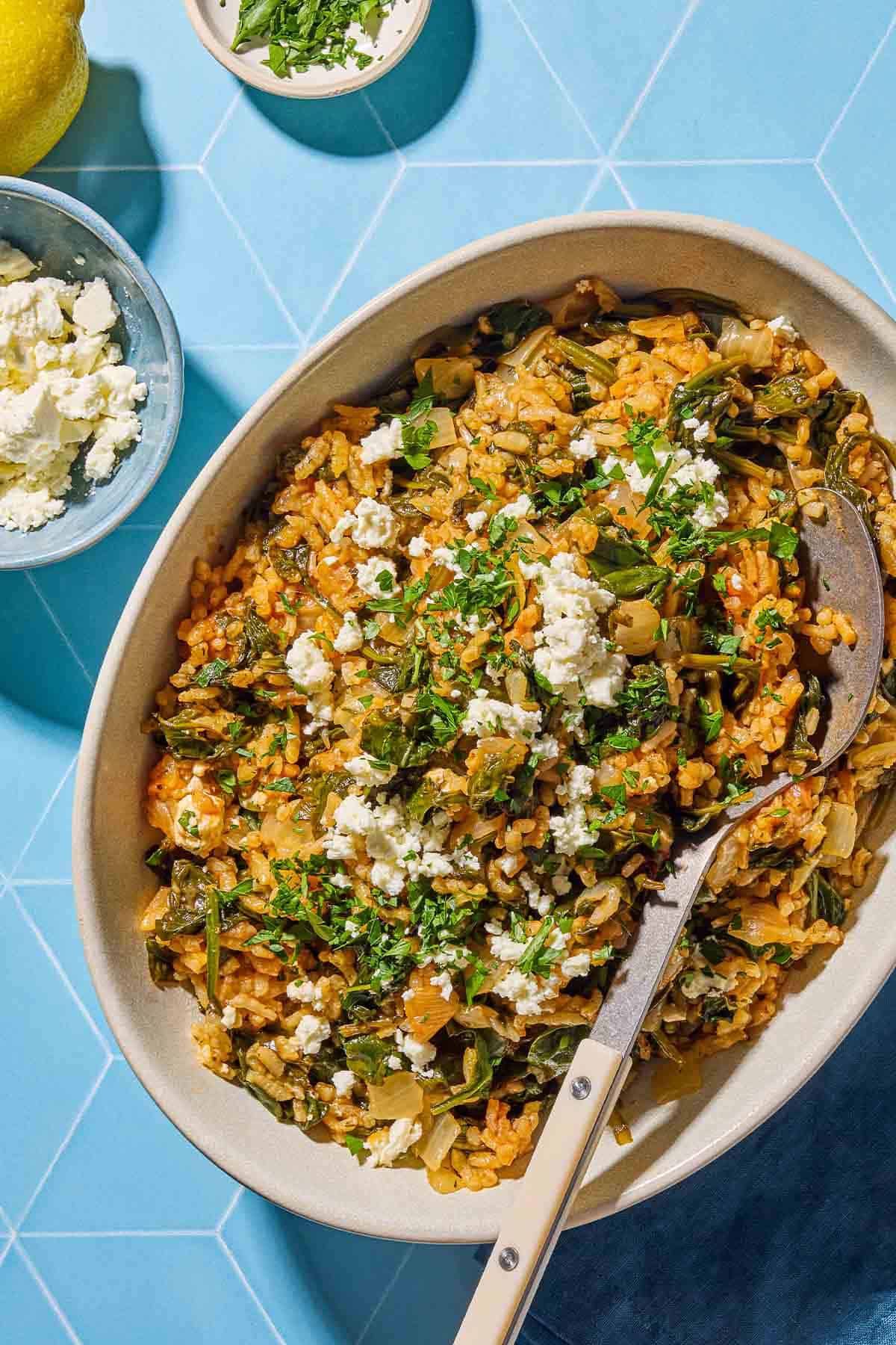 An overhead photo of spanakorizo in a serving bowl with a serving spoon. This is surrounded by a lemon and bowls of chopped parsley and feta.