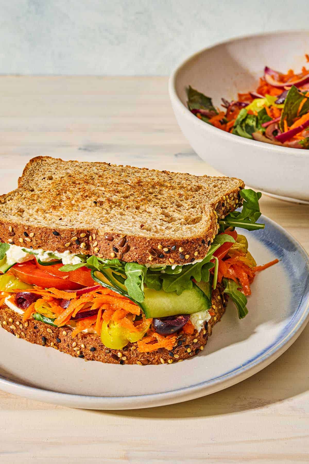A veggie sandwich on a plate in front of a bowl of the Greek-style slaw.