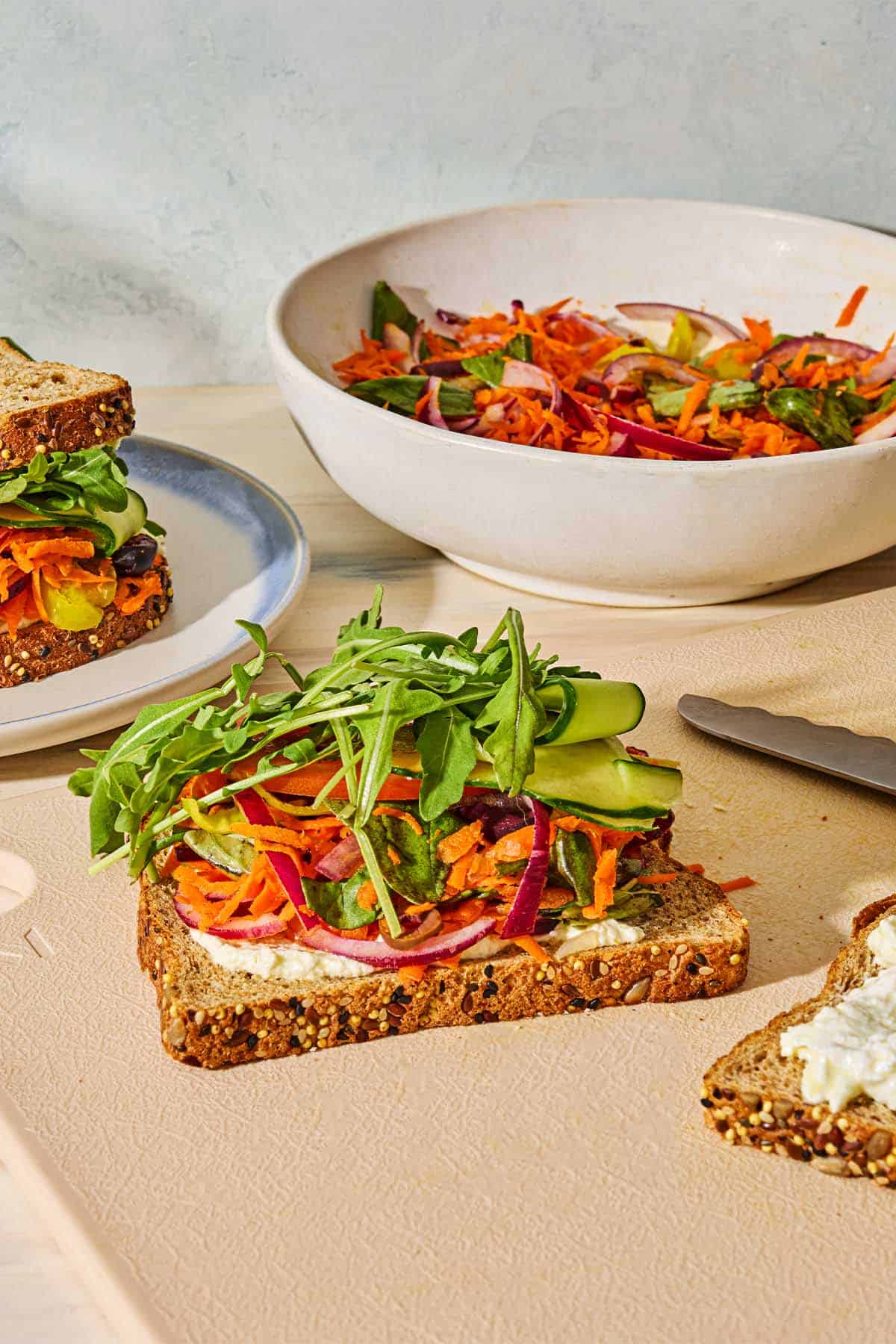 A piece of toast with the whipped feta spread and the Greek-style slaw on a cutting board with a knife. Next to this is a veggie sandwich on a plate and the rest of the slaw in a bowl.