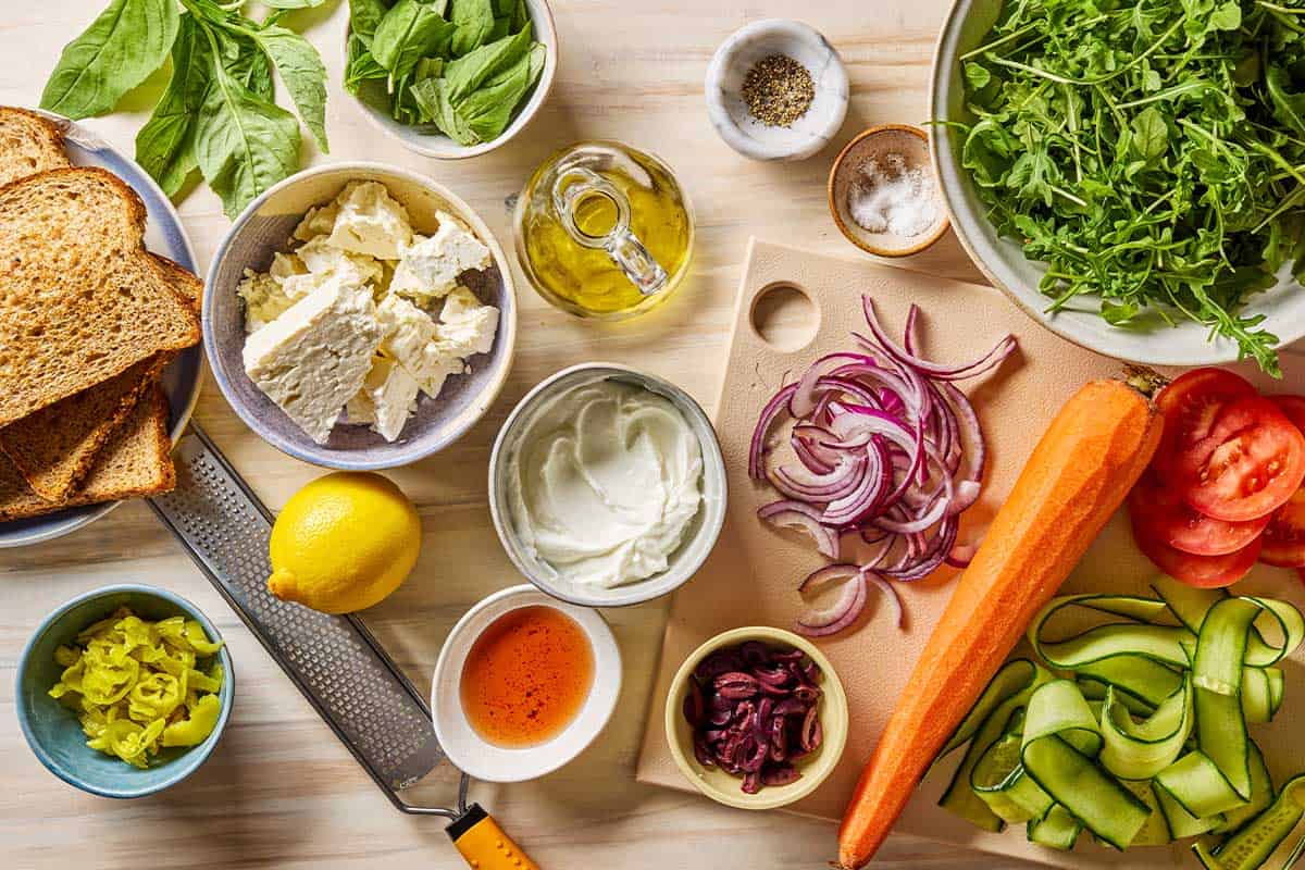 Ingredients for the veggie salad sandwich recipe including toasted bread, a carrot, red onion, peperoncini, kalamata olives, basil, red wine vinegar, olive oil, salt, pepper, cucumber, tomato, arugula, feta, greek yogurt and lemon.
