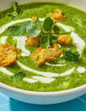 Side shot of a bowl of watercress soup drizzled with yogurt sauce and finished with croutons and mint.