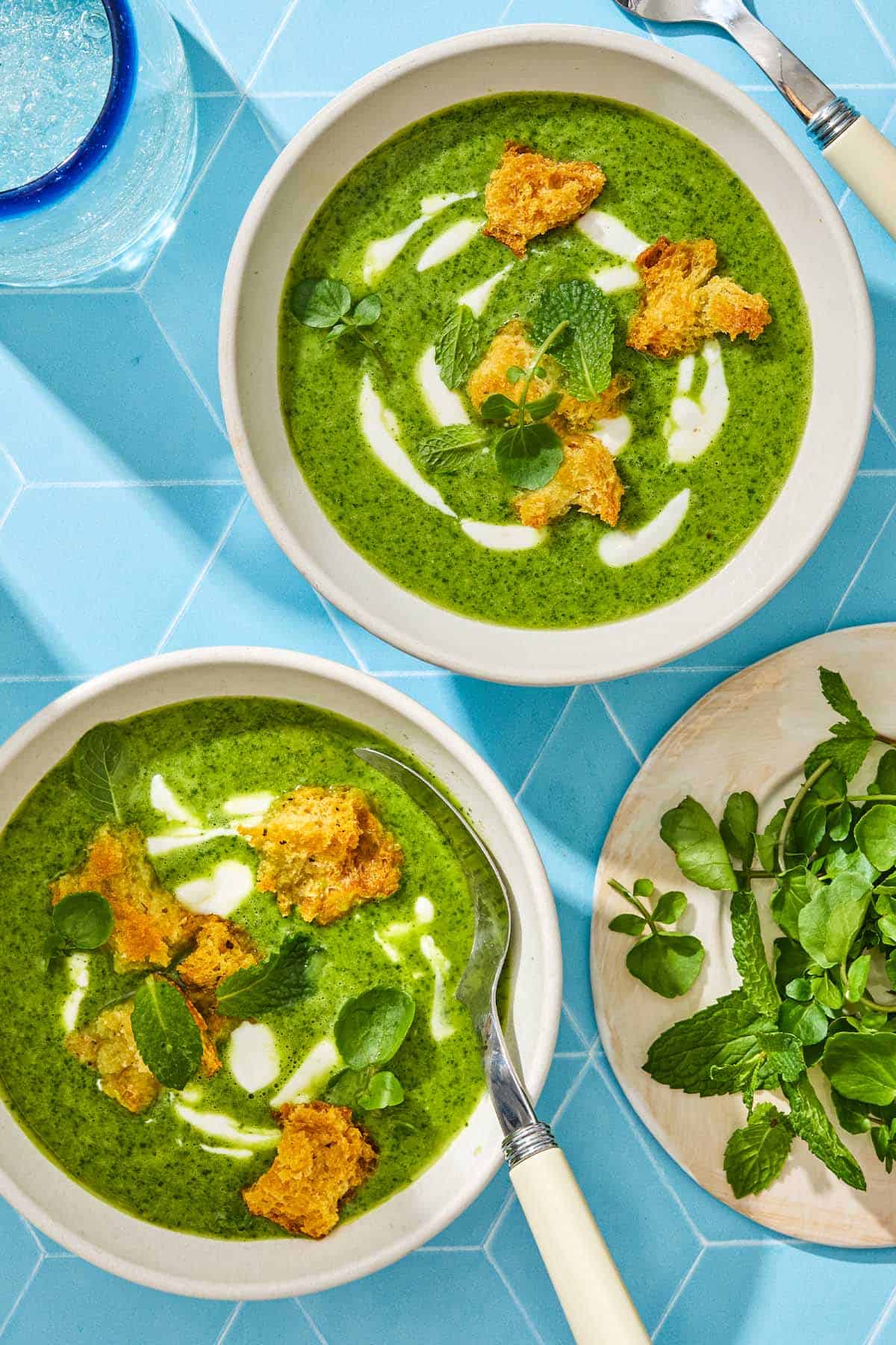 Two bowls of watercress soup with croutons, mint, and a plate with more mint on the side.