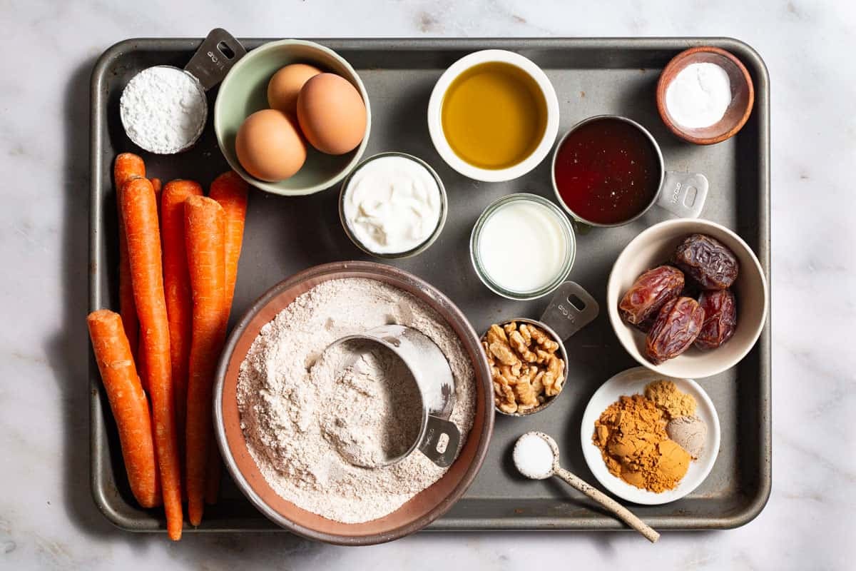 ingredients for healthy carrot cake including whole wheat flour, olive oil, greek yogurt, milk, honey, eggs, baking powder, salt, cinnamon, cardamom, ginger, carrots, dates, walnuts, and powdered sugar.