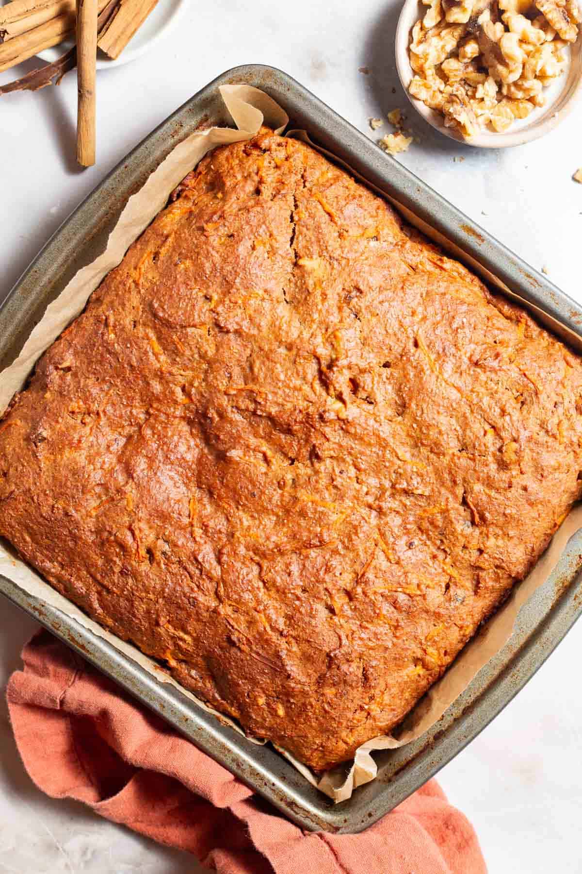 An overhead photo of a carrot cake in a parchment lined baking pan fresh out of the oven. Next to this is a kitchen towel and bowls of walnuts and cinnamon sticks.