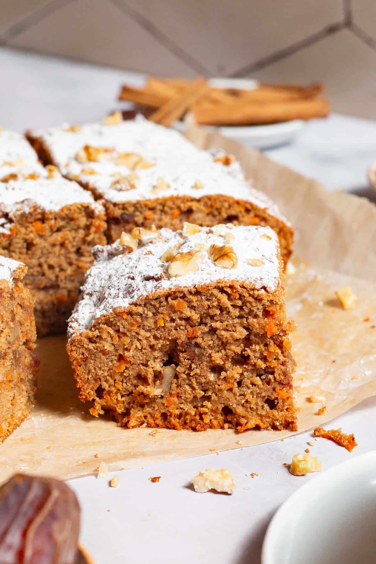 A close up of a slice of the healthy carrot cake in front of the rest of the cake on a sheet of parchment paper.
