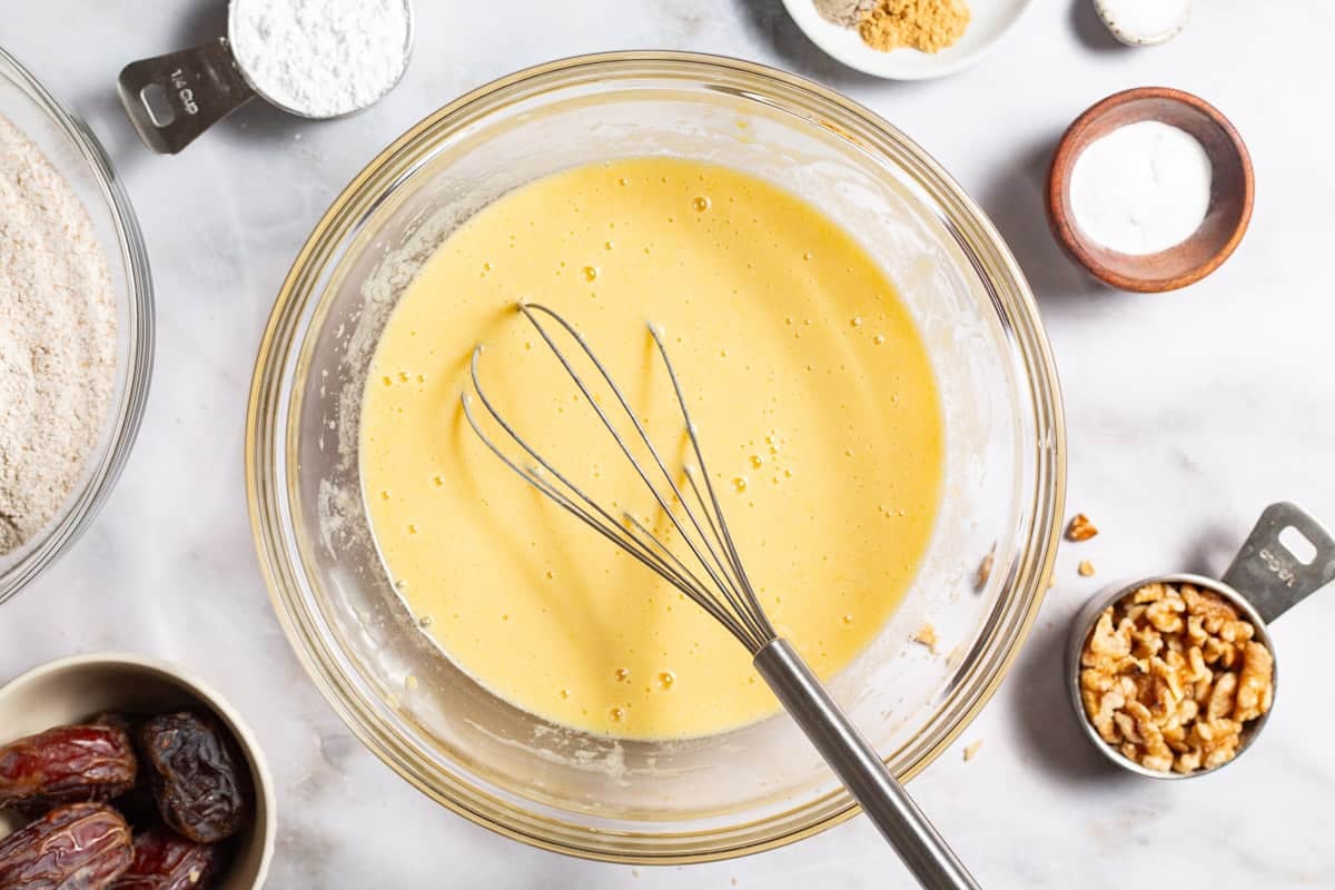 The wet ingredients for the healthy carrot cake mixed together in a bowl with a whisk. Surrounding this are bowls of dates, spices, baking powder and the dry ingredients for the dough, and measuring cups of walnuts and powdered sugar.