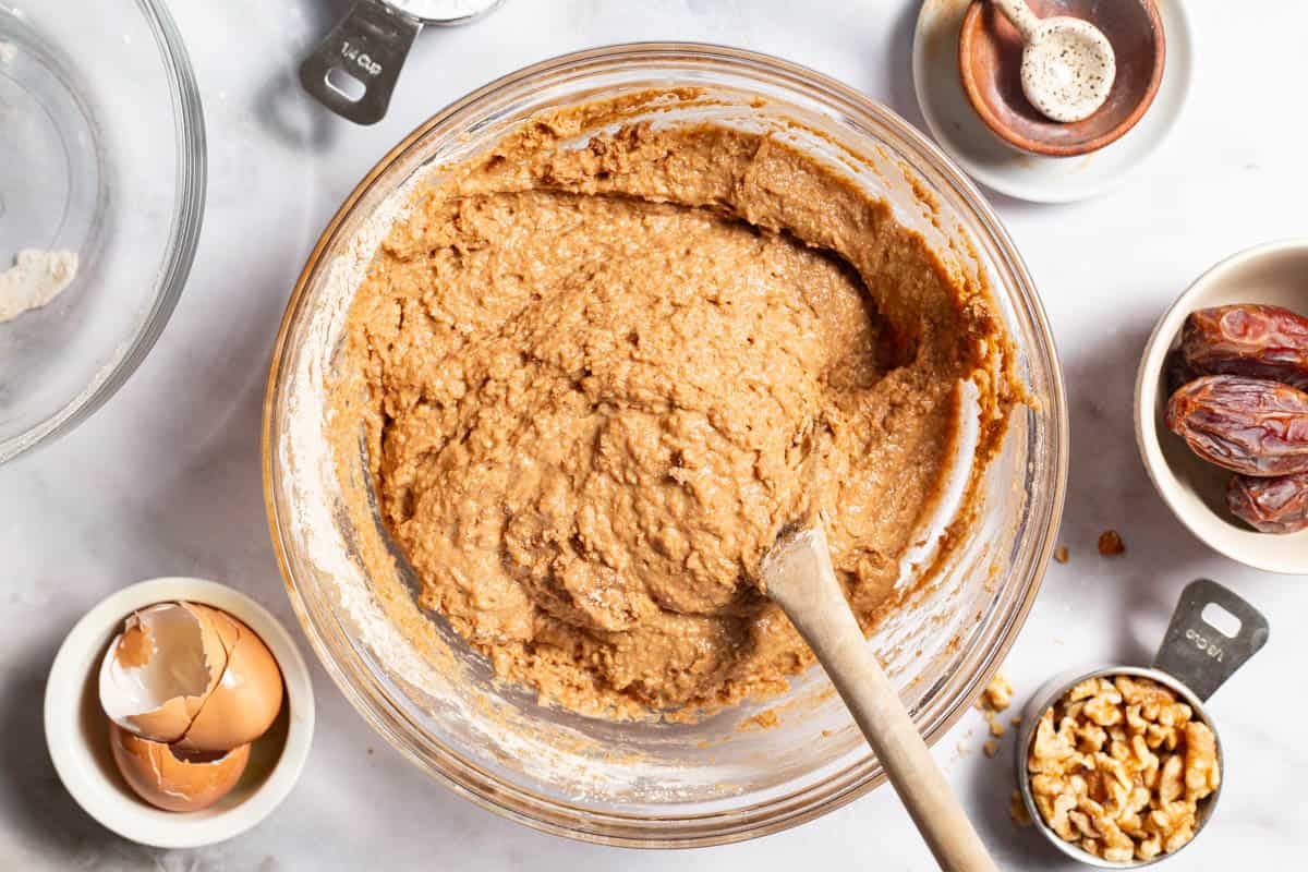 The ingredients for the healthy carrot cake in a bowl with a wooden spoon just after being mixed together. Next to this is a bowl of egg shells, a measuring cup with walnuts, a bowl of dates, and 3 other empty bowls, on with a spoon.