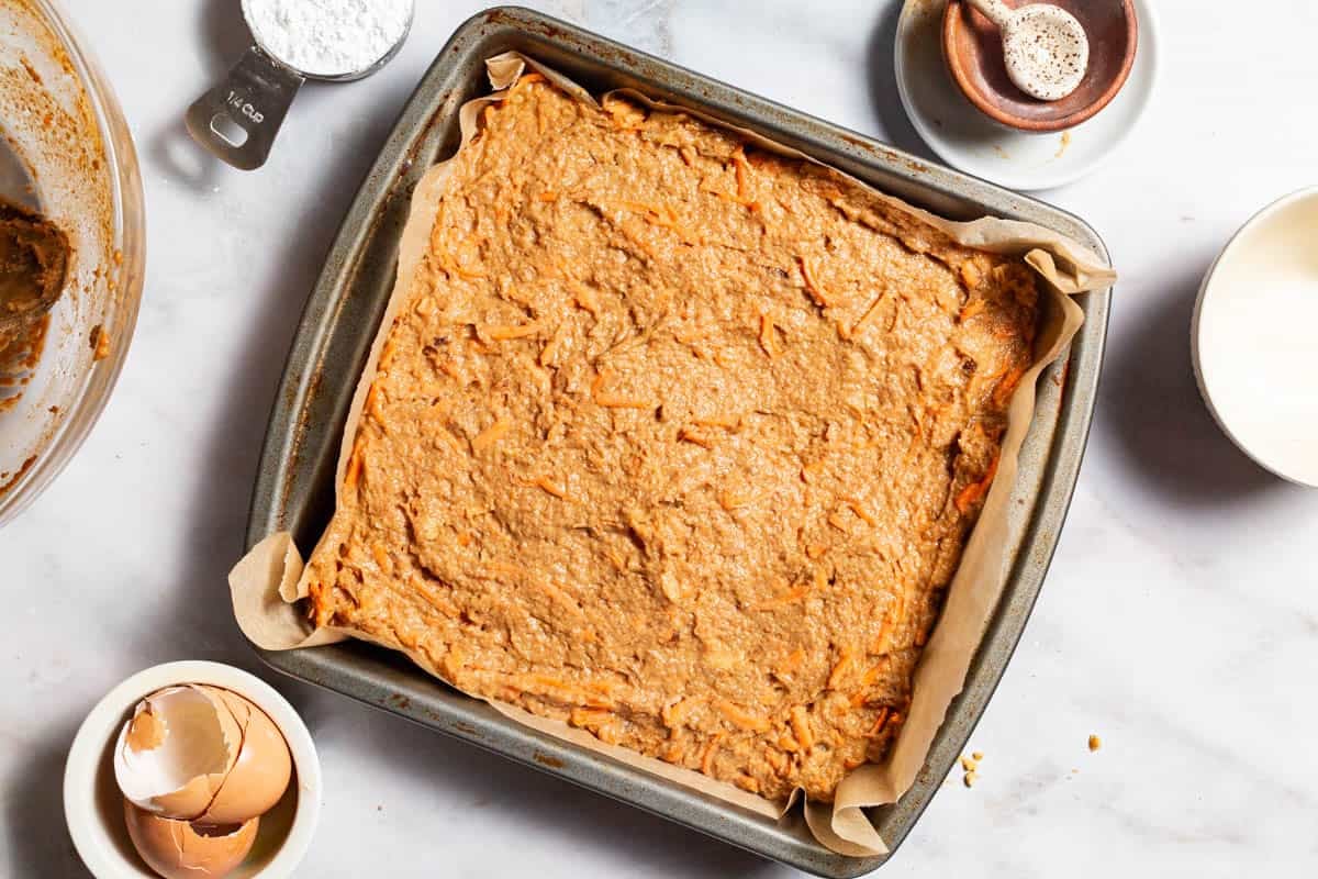 An overhead photo of the healthy carrot cake batter poured into a parchment lined baking dish. Next to this is a measuring cup with powdered sugar, 4 empty bowls and bowl of egg shells.