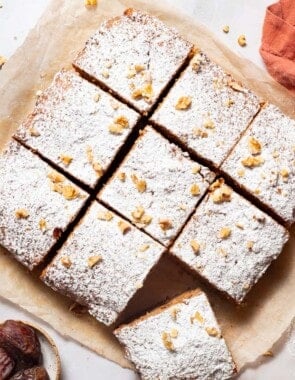An and overhead photo of 9 slices of healthy carrot cake on a sheet of parchment paper. Next to these are bowls of walnuts, cinnamon sticks and dates, a sieve with powdered sugar, and a kitchen towel.