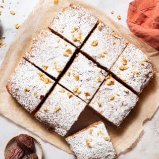 An and overhead photo of 9 slices of healthy carrot cake on a sheet of parchment paper. Next to these are bowls of walnuts, cinnamon sticks and dates, a sieve with powdered sugar, and a kitchen towel.