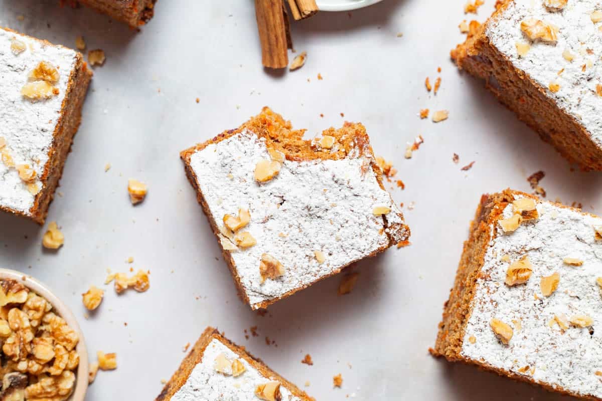 An and overhead close up photo of 5 slices of healthy carrot cake, one with a bite taken out of it. Next to this is a bowl of walnuts.