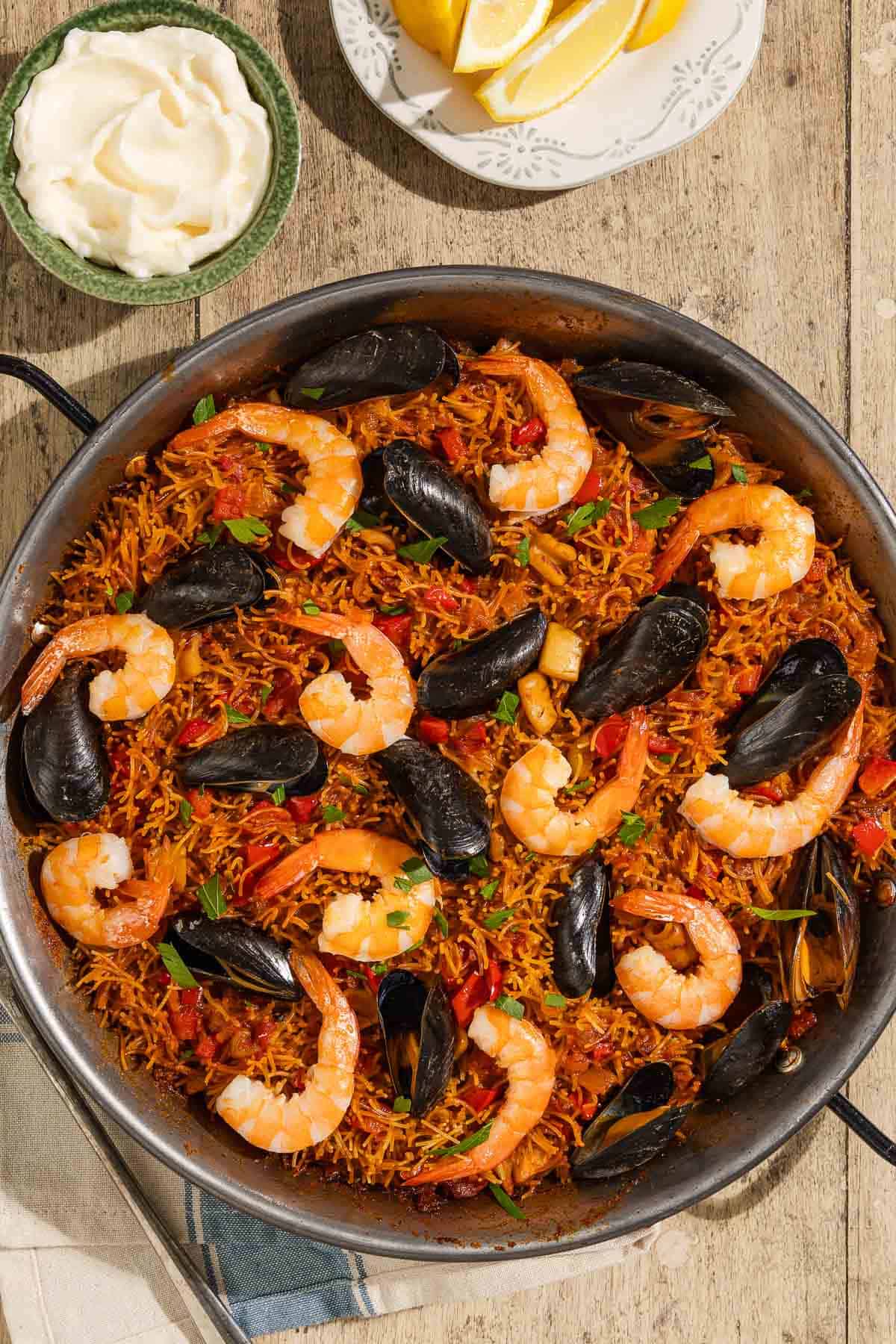 An overhead photo of Catalan fideua in a skillet surrounded by a bowl of aioli, lemon wedges, and a kitchen towel.