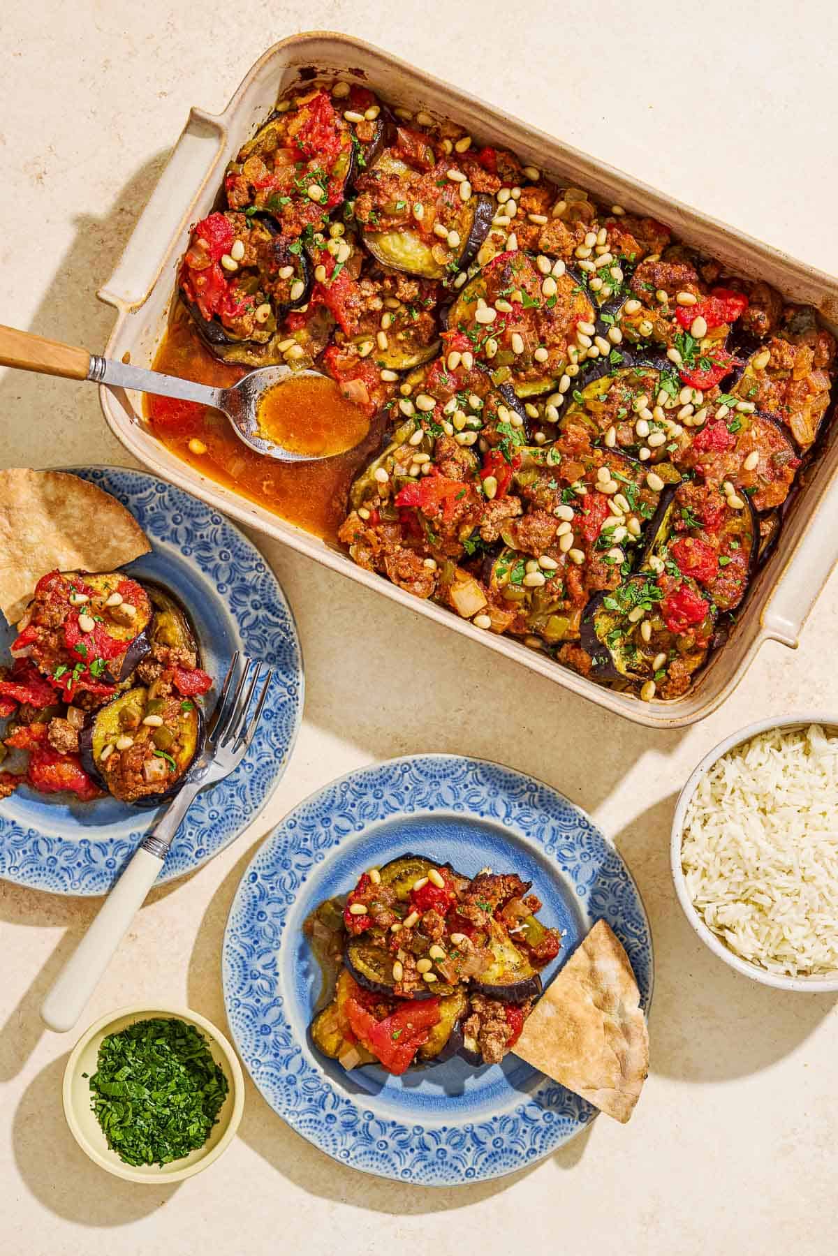 An overhead photo of 2 servings of egyptian moussaka on plates, each with a slice of pita bread and one with a fork. Next to these is the rest of the moussaka in a baking dish with a spoon and bowls of rice and chopped parsley.