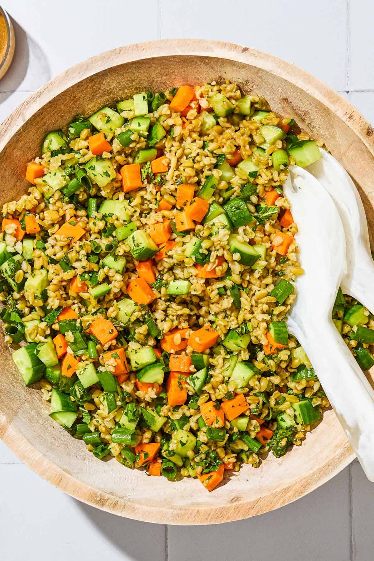 An overhead close up of herby freekeh salad in a serving bowl with serving utensils.