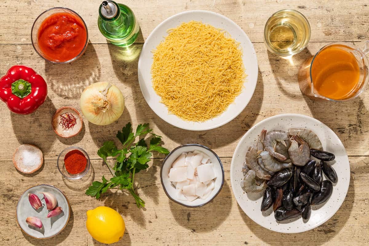Ingredients for Catalan fideua including fideos, seafood stock, olive oil, shrimp, squid, mussels, onion, red bell pepper, garlic, canned crushed tomatoes, smoked paprika, salt, saffron, white wine, parsley, and lemon.