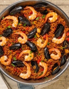 An overhead photo of Catalan fideua in a skillet surrounded by a bowl of aioli, lemon wedges, a kitchen towel and a serving spoon.