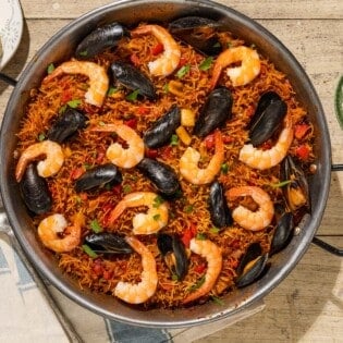 An overhead photo of Catalan fideua in a skillet surrounded by a bowl of aioli, lemon wedges, a kitchen towel and a serving spoon.