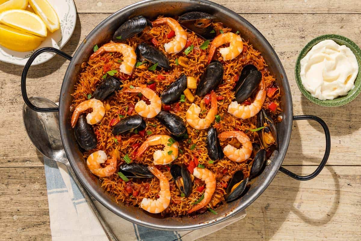 An overhead photo of Catalan fideua in a skillet surrounded by a bowl of aioli, lemon wedges, a kitchen towel and a serving spoon.