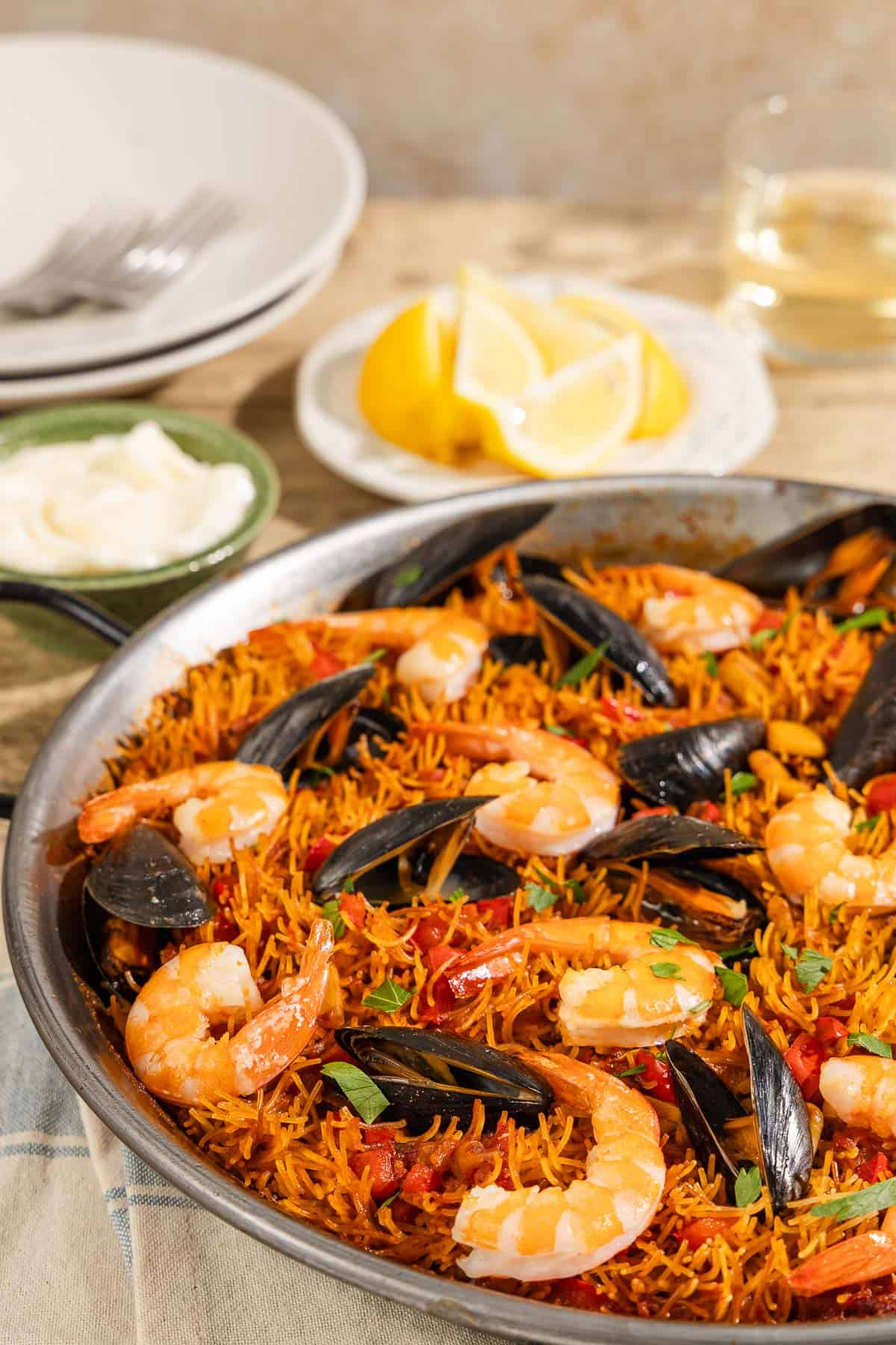 A close up of Catalan fideua in a skillet in front of a bowl of aioli, lemon wedges, a glass of wine, 2 plates and 2 forks.