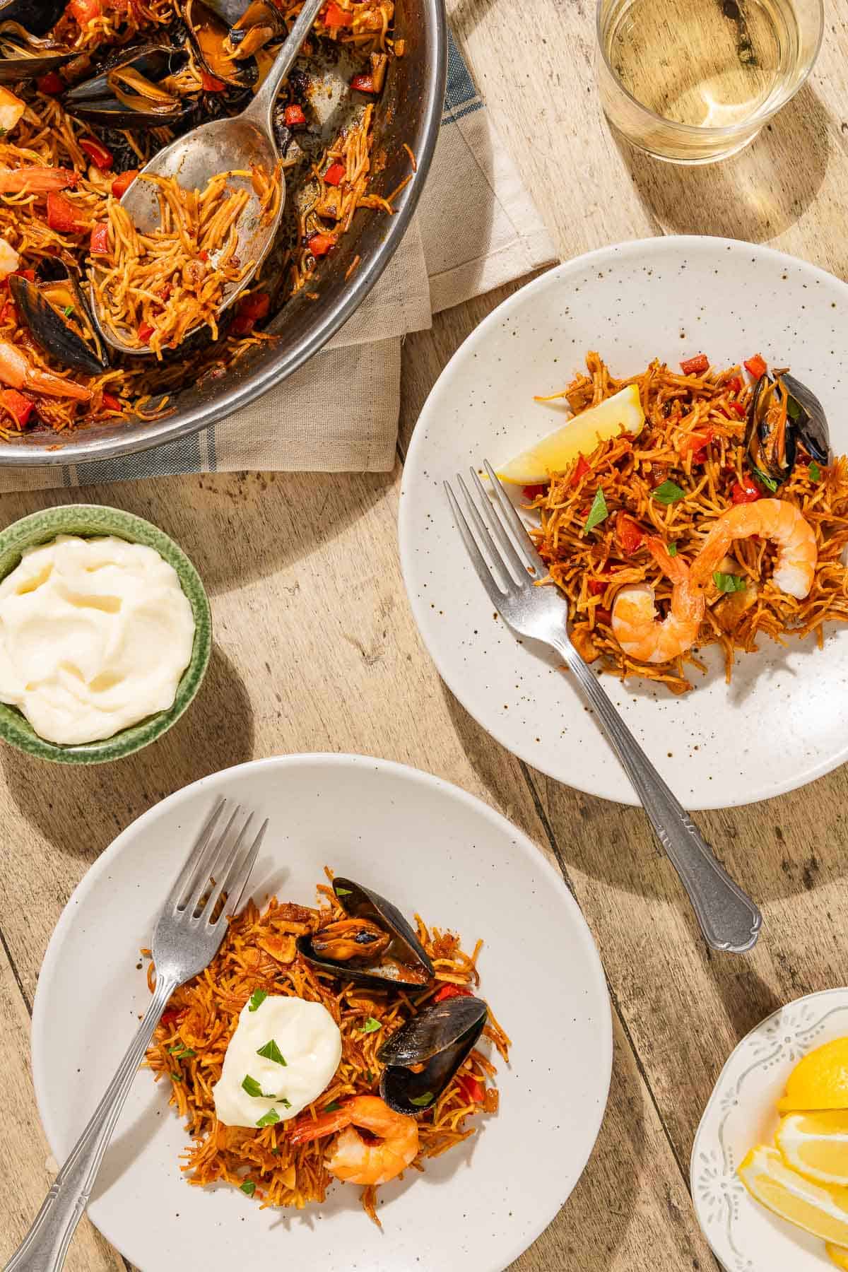 An overhead photo of two servings of catalan fideua on plates with forks. Next to these is a glass of wine, a bowl of aioli, lemon wedges, and the rest of the fideua in a skillet with a serving spoon.