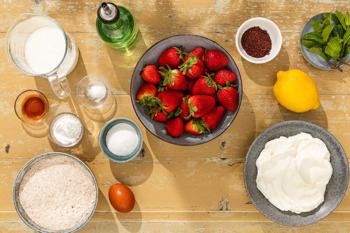 Ingredients for strawberry shortcake including strawberries, sumac, lemon, sugar, flour, baking powder, salt, milk, olive oil, vanilla extract, an egg, water, labneh, and mint.