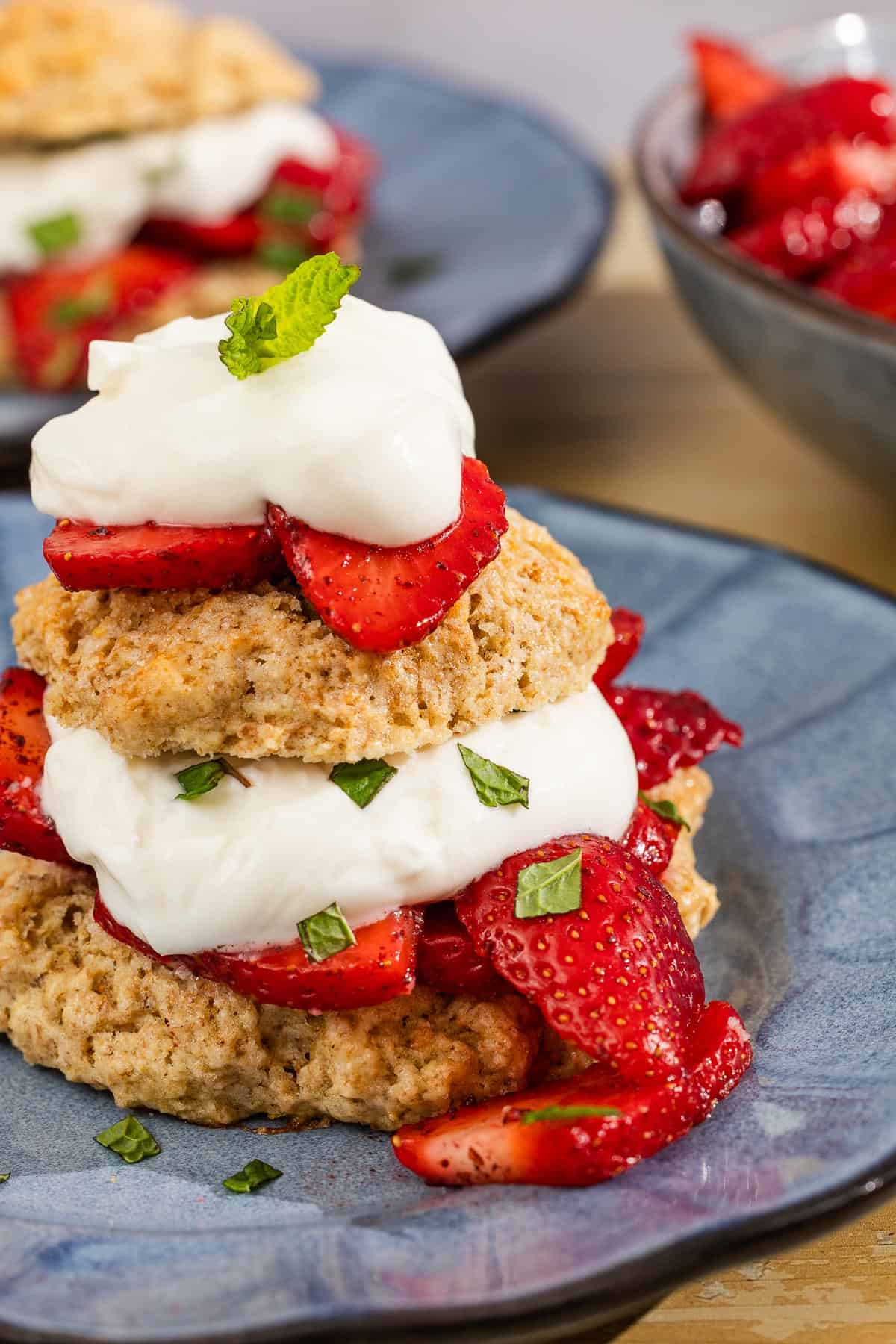 A close up of a strawberry shortcake on a plate in front of another shortcake on a plate and a bowl of the strawberries.