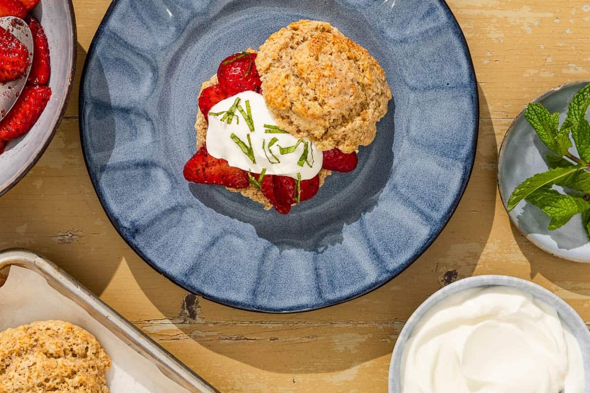 An overhead photo of a strawberry shortcake on a blue plate. Next to this is a bowl of whipped cream, a small plate of mint, a bowl of strawberries with a spoon, and a baked shortcake on a parchment-lined baking sheet.