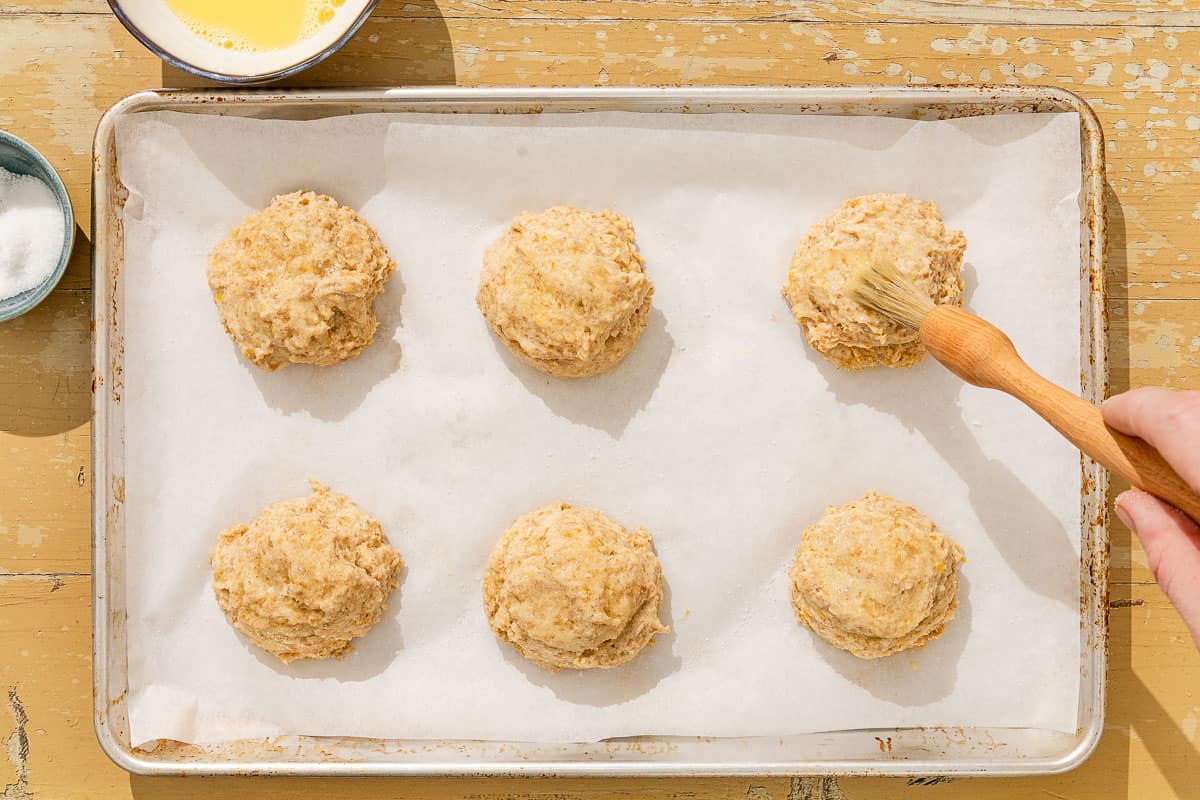 Six unbaked shortcakes on a parchment-lined baking sheet with one being brushed with an egg wash. Next to this is a bowl of the egg wash and a small bowl of sugar.