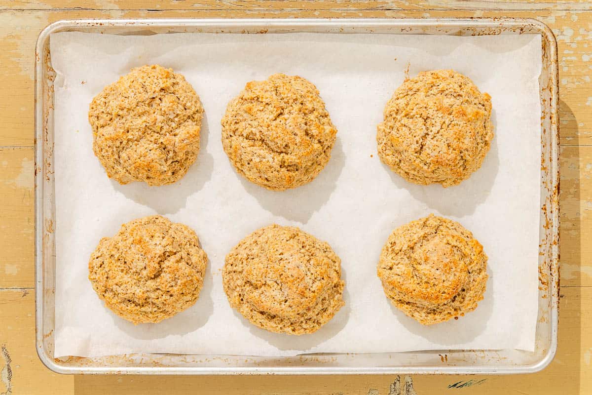 Six baked shortcakes on a parchment-lined baking sheet with one being brushed with an egg wash. Next to this is a bowl of the egg wash and a small bowl of sugar.