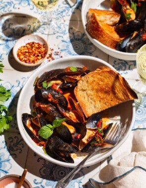 Mussels marinara in a bowl with a piece of toasted bread and a fork. Next to this is another serving of mussels, bowls of salt and red pepper flakes, two glasses of wine and a cloth napkin.