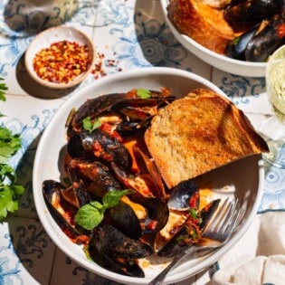 Mussels marinara in a bowl with a piece of toasted bread and a fork. Next to this is another serving of mussels, bowls of salt and red pepper flakes, two glasses of wine and a cloth napkin.