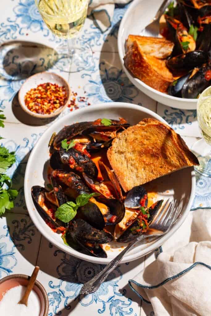 Mussels marinara in a bowl with a piece of toasted bread and a fork. Next to this is another serving of mussels, bowls of salt and red pepper flakes, two glasses of wine and a cloth napkin.