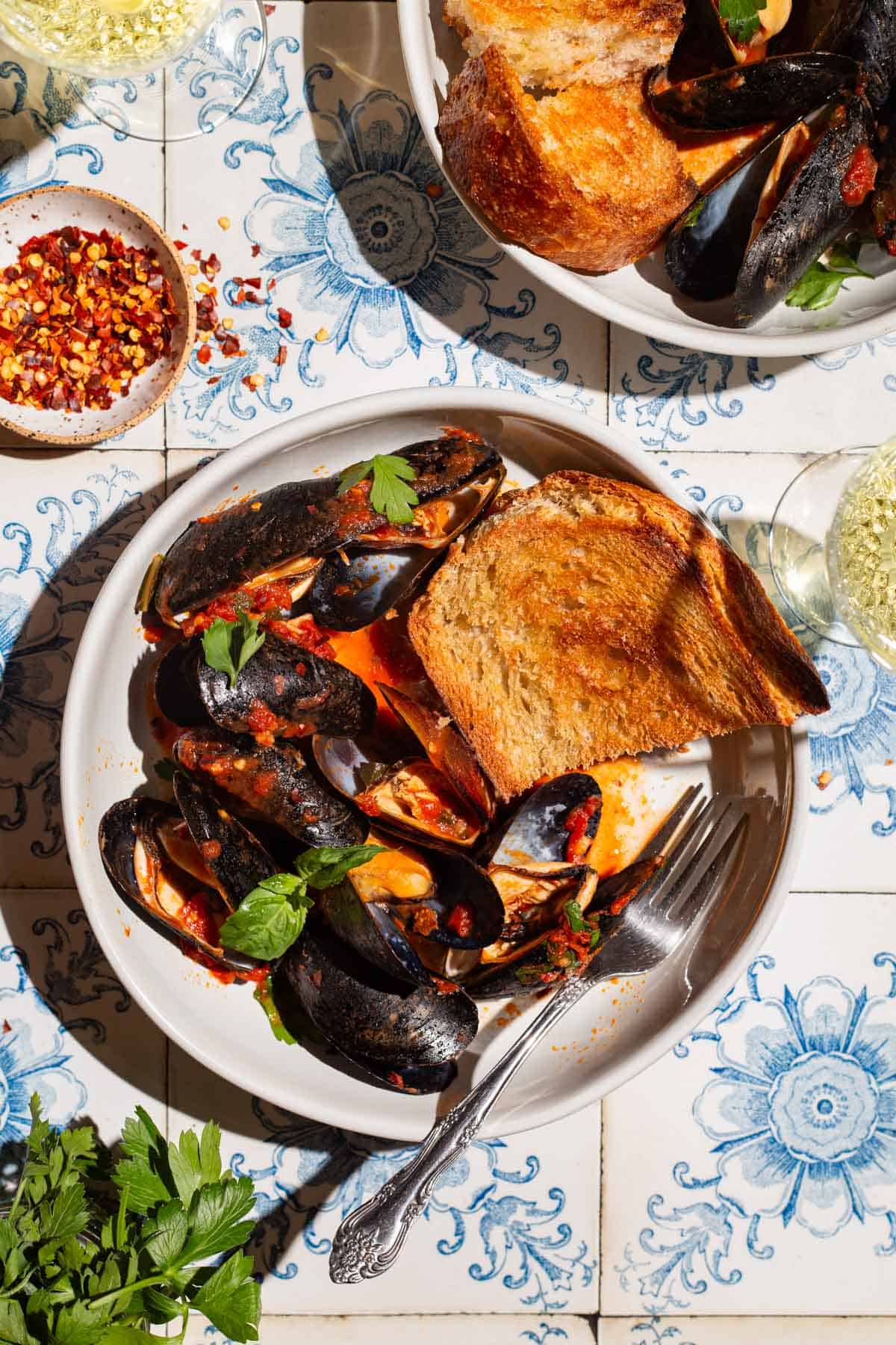 An overhead photo of mussels marinara in a bowl with a piece of toasted bread and a fork. Next to this is another serving of mussels, a bowl of red pepper flakes, parsley, and two glasses.