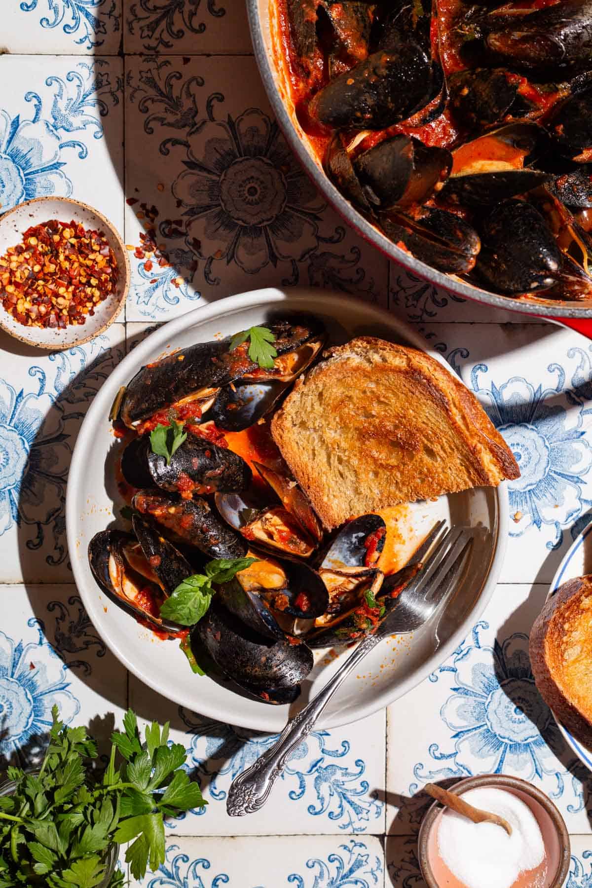 An overhead photo of mussels marinara in a bowl with a piece of toasted bread and a fork. Next to this is a pot of mussels, bowls of salt and red pepper flakes and parsley.