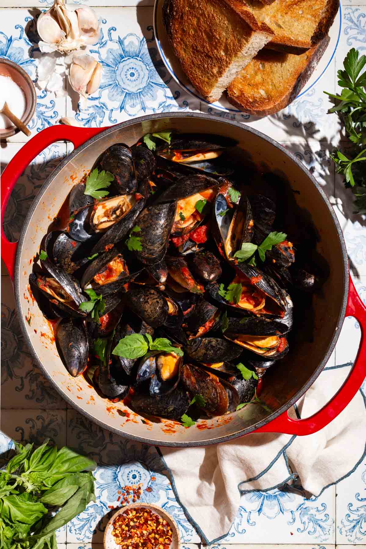 An overhead photo of a pot of mussels marinara surrounded by garlic cloves, bowls of salt and red pepper flakes, a plate of toasted bread, basil and a cloth napkin.