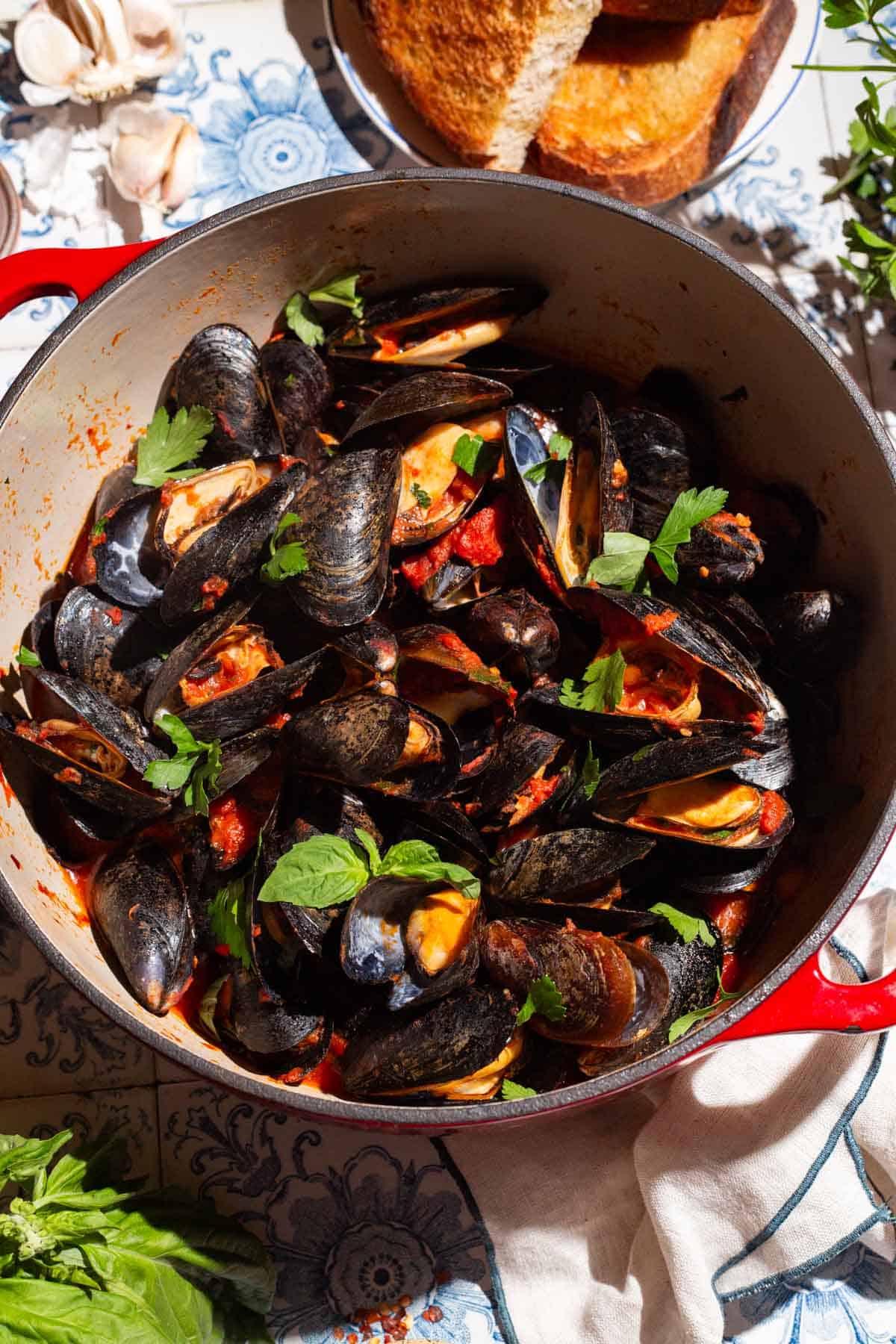 An overhead photo of a pot of mussels marinara surrounded by garlic cloves, a plate of toasted bread, basil and a cloth napkin.