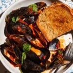 An overhead photo of mussels marinara in a bowl with a piece of toasted bread and a fork.